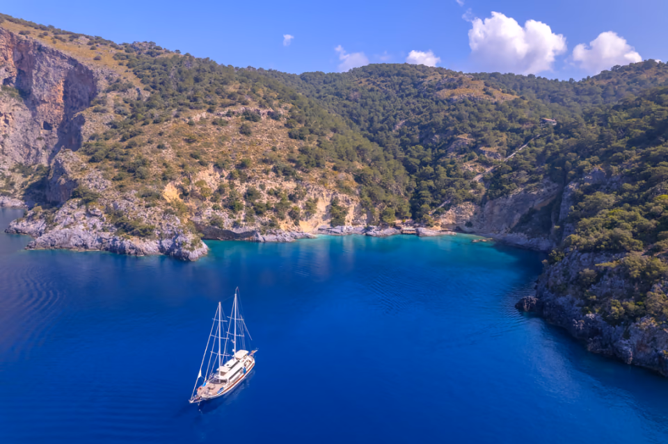 A white gulet at anchor in a deep blue bay ringed by forested hills and limestone crags.