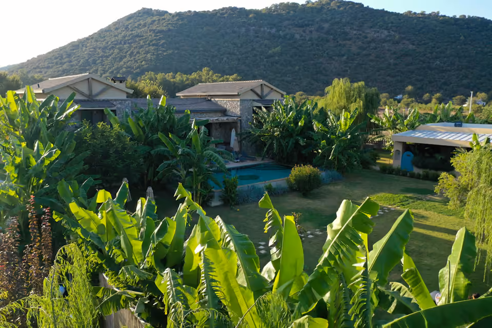 The villa and its garden in the Kayaköy valley at golden hour: stone buildings and a pool among banana palms, the wooded ridge rising behind.
